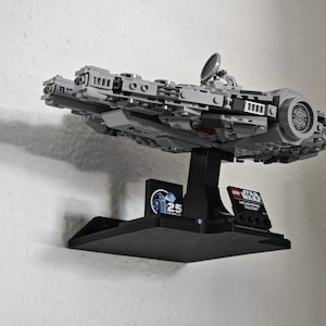 以下が含まれることがあります： 黒い壁掛けに、グレーとブラックのLEGOミレニアム・ファルコンのモデルが飾られています。モデルは、LEGOスター・ウォーズミレニアム・ファルコン25周年記念の小さなロゴが付いた黒いスタンドに載せられています。