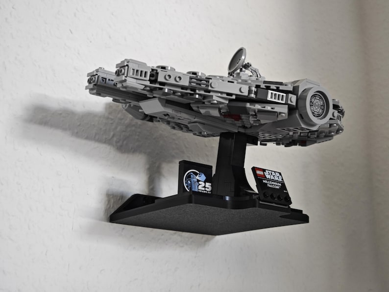 以下が含まれることがあります： 黒い壁掛けに、グレーとブラックのLEGOミレニアム・ファルコンのモデルが飾られています。モデルは、LEGOスター・ウォーズミレニアム・ファルコン25周年記念の小さなロゴが付いた黒いスタンドに載せられています。