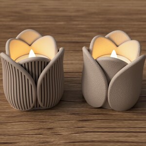 May include: Two taupe-colored tulip-shaped candle holders with lit candles inside. One holder has vertical lines, the other has a smooth texture. The candle holders sit on a wooden surface.