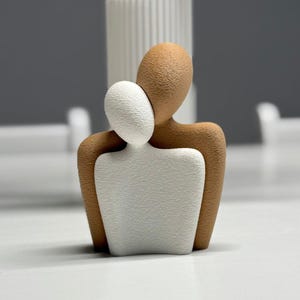 May include: A contemporary sculpture of two embracing abstract figures. One figure is white, the other is brown, with a textured finish. The sculpture is set against a neutral backdrop, suggesting a minimalist design.