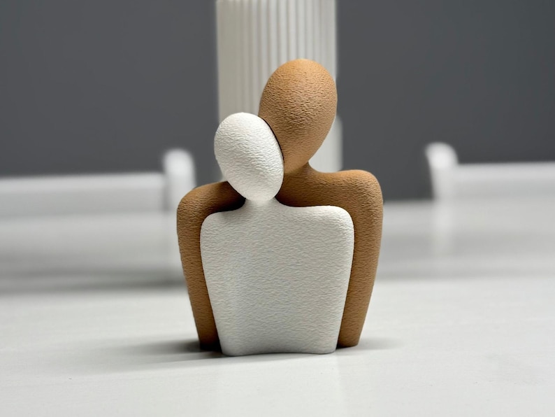 May include: A contemporary sculpture of two embracing abstract figures. One figure is white, the other is brown, with a textured finish. The sculpture is set against a neutral backdrop, suggesting a minimalist design.