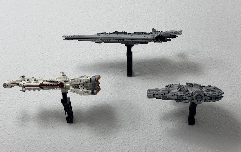 May include: Three Star Wars spaceship models, including the Millennium Falcon, a Star Destroyer, and a smaller ship, are displayed on black stands against a white wall. The models are detailed and grey.