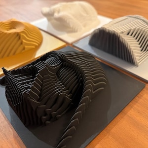 Puede incluir: Cuatro esculturas impresas en 3D, cada una con un color y un dise&ntilde;o diferente. Las esculturas est&aacute;n hechas de cart&oacute;n en capas y est&aacute;n dispuestas sobre una superficie de madera.