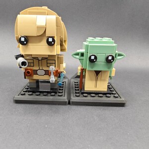 Może przedstawiać: Dwie minifigurki Lego, jedna przedstawiająca Luke'a Skywalkera, a druga Yodę, stoją na czarnych podstawkach. Luke jest ubrany w brązowy str&oacute;j Jedi i trzyma miecz świetlny. Yoda jest ubrany w brązową szatę i trzyma miecz świetlny.