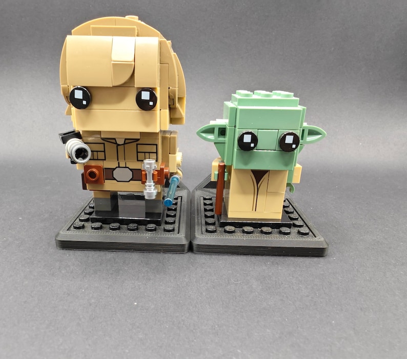 Może przedstawiać: Dwie minifigurki Lego, jedna przedstawiająca Luke'a Skywalkera, a druga Yodę, stoją na czarnych podstawkach. Luke jest ubrany w brązowy str&oacute;j Jedi i trzyma miecz świetlny. Yoda jest ubrany w brązową szatę i trzyma miecz świetlny.