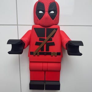 May include: A red and black 3D printed Lego figurine of Deadpool. The figurine has a black mask with white eyes and a red and black suit with a brown belt.