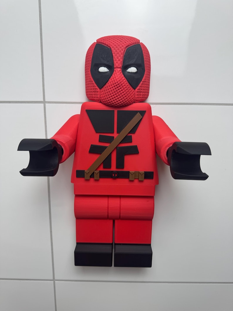 May include: A red and black 3D printed Lego figurine of Deadpool. The figurine has a black mask with white eyes and a red and black suit with a brown belt.