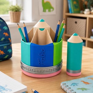May include: A multi-compartment pencil holder shaped like a large pencil with a teal, blue, and green body, a silver metal-look band, and a pink base. The name Fabio is embossed on the blue section. A matching smaller pencil-shaped container sits nearby.