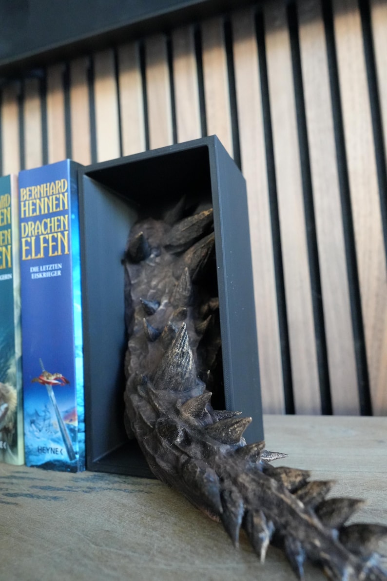May include: A black bookend with a dragon tail design. The tail is bronze-colored with sharp spikes. The bookend is holding up a row of books with blue and white covers. The books are on a wooden surface.