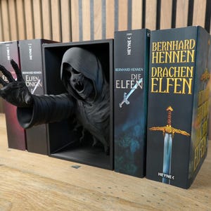 May include: A gothic book display with a dark, hooded figure reaching out from a black box. The display is accompanied by German-language books, including "Die Elfen" and "Drachenelfen", creating a fantasy theme.
