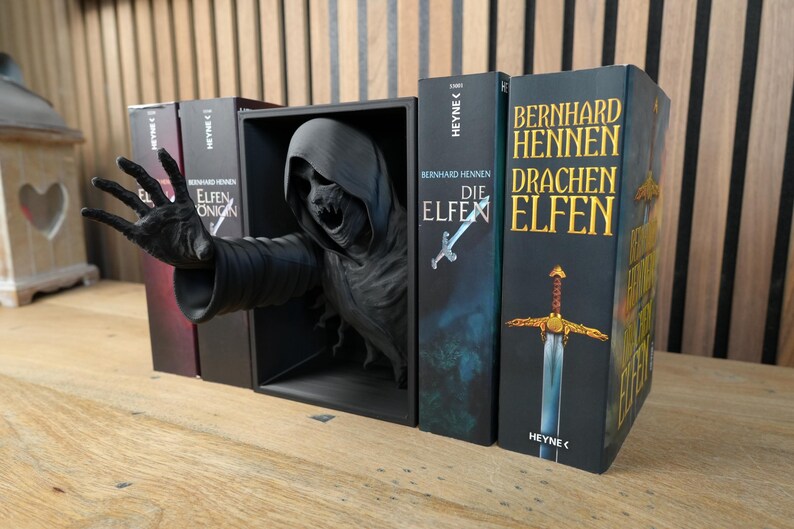 May include: A gothic book display with a dark, hooded figure reaching out from a black box. The display is accompanied by German-language books, including "Die Elfen" and "Drachenelfen", creating a fantasy theme.