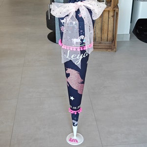 Puede incluir: Un cono escolar azul y blanco con tem&aacute;tica de unicornio con pompones rosas y una base blanca. El cono est&aacute; decorado con un lazo de tul blanco y el nombre "Leya" est&aacute; escrito en el cono.