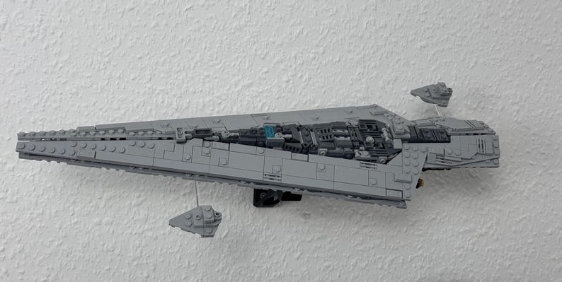 May include: A grey Lego model of a Star Destroyer spaceship, mounted on a white wall. The model is detailed with various grey blocks and features two smaller Lego ships. The model is approximately 61 cm long.