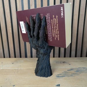 May include: A black, stylized hand-shaped bookend holding a book with a burgundy cover. The bookend has a textured, tree-like base. The book's title is partially visible, with the word "HEYNE" printed on the spine.