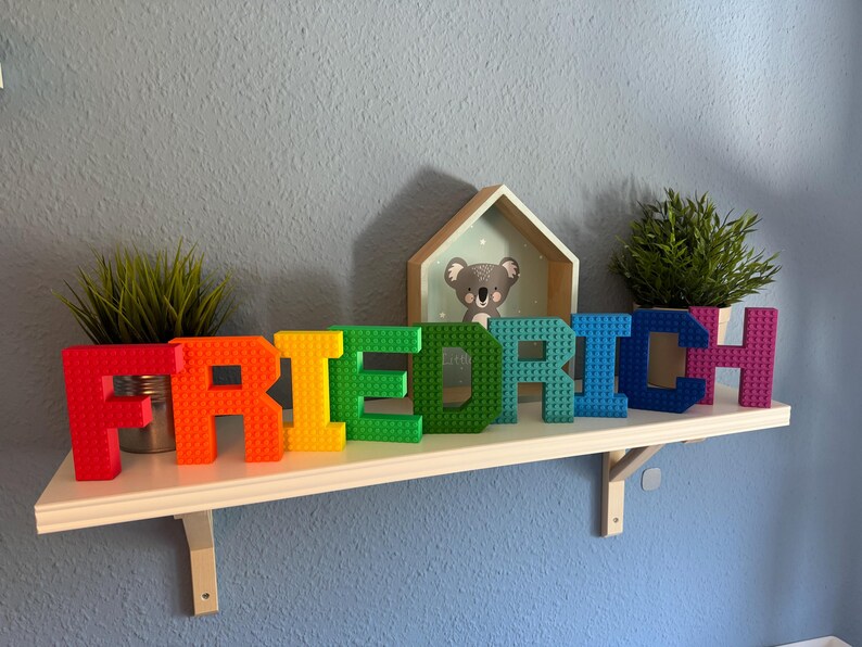 May include: A colorful name sign spelling "FRIEDRICH" in large, block-style letters. The letters are in a rainbow of colors and sit on a white shelf with small potted plants and a koala bear picture.