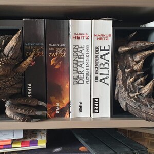May include: A detailed dragon head and claw bookend set in shades of brown and gold, placed on a bookshelf with books. The bookends are designed to appear as if the dragon is emerging from the books. The books have titles in German.