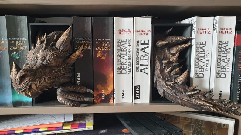 May include: A detailed dragon head and claw bookend set in shades of brown and gold, placed on a bookshelf with books. The bookends are designed to appear as if the dragon is emerging from the books. The books have titles in German.