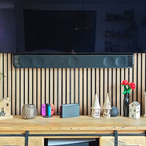 May include: A wooden console table with various decorative items, including a small house-shaped sign that says "Home", a candle holder, and a vase with red flowers. A black soundbar is mounted below a large TV.