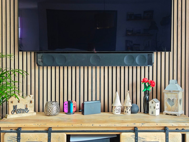 May include: A wooden console table with various decorative items, including a small house-shaped sign that says "Home", a candle holder, and a vase with red flowers. A black soundbar is mounted below a large TV.