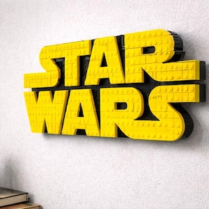 Pode incluir: Um logotipo "STAR WARS" amarelo e preto feito de blocos interligados, montado em uma parede branca. As letras s&atilde;o amarelas e o fundo &eacute; preto. Uma pequena planta em vaso est&aacute; localizada abaixo do logotipo.