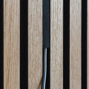 Op de afbeelding: Close-up van een houten paneel met verticale, afwisselende lichtbruine en zwarte strepen. Een grijze kabel loopt verticaal door het paneel. De houtnerf is zichtbaar in de lichtbruine secties.