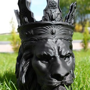 May include: A black lion head planter with a crown. The planter is made of a dark material and sits in green grass. The lion has a detailed mane and a stern expression. The crown has ornate details.