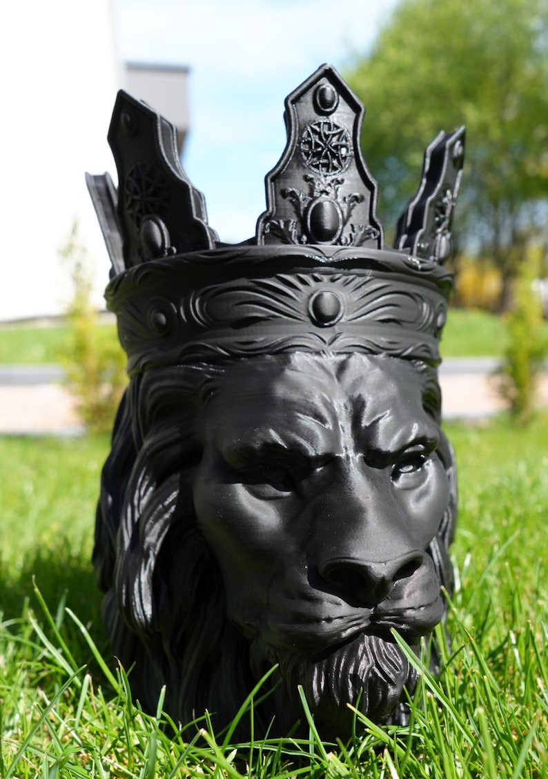 May include: A black lion head planter with a crown. The planter is made of a dark material and sits in green grass. The lion has a detailed mane and a stern expression. The crown has ornate details.