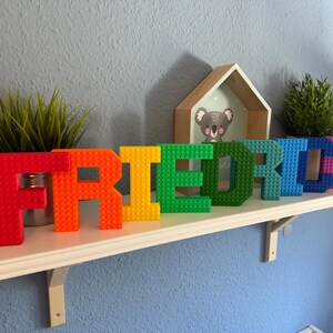 May include: A colorful, rainbow-hued name, "FRIEDRICH", constructed from interlocking building blocks, sits on a white shelf. The letters are red, orange, yellow, green, blue, and pink. Small potted plants and a koala picture frame are also on the shelf.