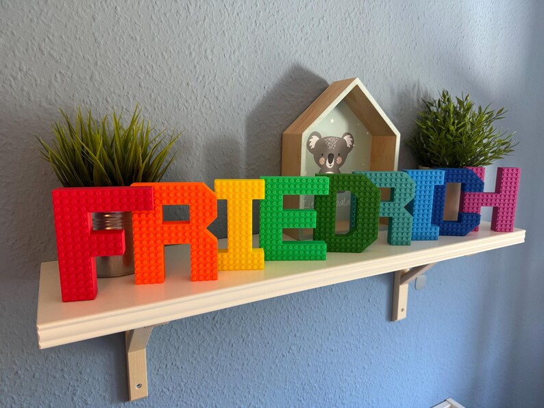 May include: A colorful, rainbow-hued name, "FRIEDRICH", constructed from interlocking building blocks, sits on a white shelf. The letters are red, orange, yellow, green, blue, and pink. Small potted plants and a koala picture frame are also on the shelf.