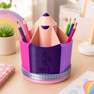 May include: A multi-colored pencil-shaped desk organizer featuring a central compartment and several outer slots. The exterior is decorated with vertical panels in shades of pink, purple, and dark violet, with the name Leya embossed on the side.