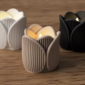 May include: Three tulip-shaped candle holders in white, beige, and black. Each holder has a ribbed design and contains a lit candle, casting a warm glow. The candle holders are arranged on a wooden surface.