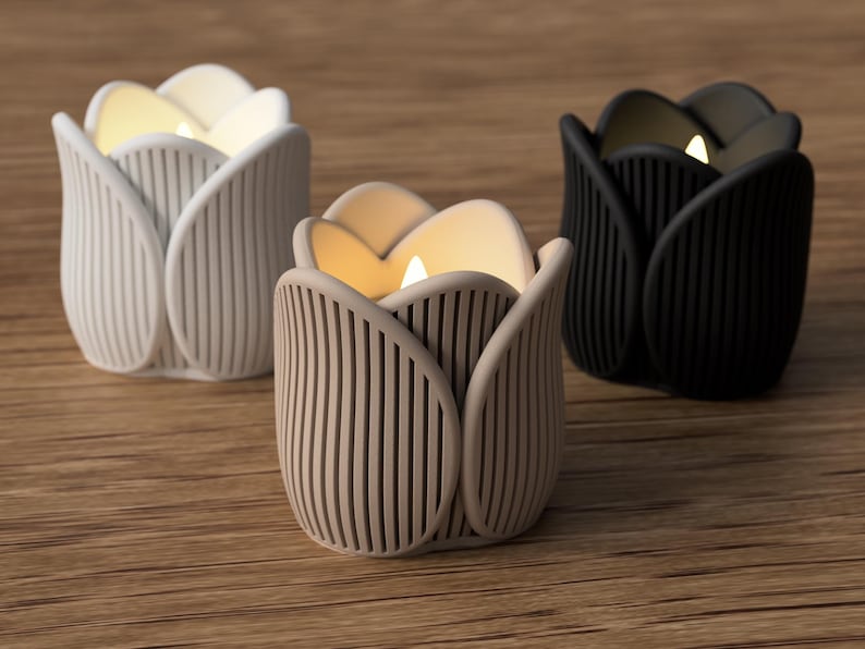 May include: Three tulip-shaped candle holders in white, beige, and black. Each holder has a ribbed design and contains a lit candle, casting a warm glow. The candle holders are arranged on a wooden surface.