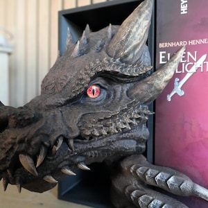 May include: A detailed, dark gray dragon head bookend with a red eye and sharp teeth. The dragon's head is emerging from a black shelf, with a book titled "Elven Light" visible. The bookend adds a fantasy theme to any bookshelf.