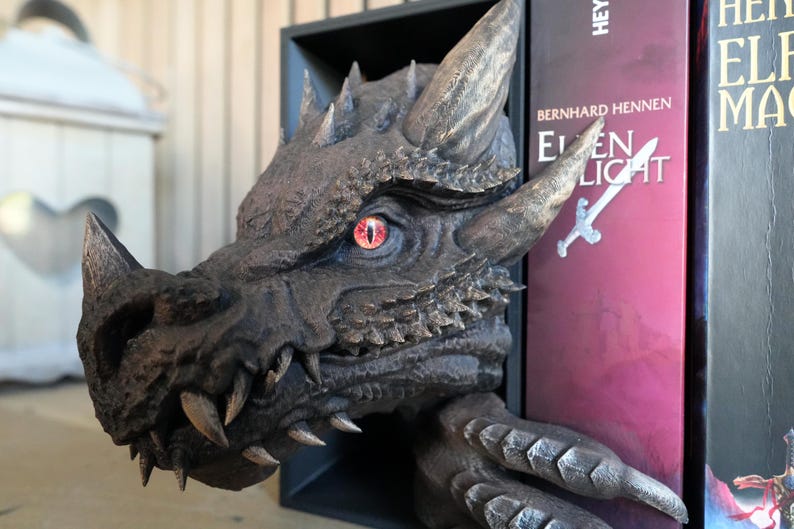 May include: A detailed, dark gray dragon head bookend with a red eye and sharp teeth. The dragon's head is emerging from a black shelf, with a book titled "Elven Light" visible. The bookend adds a fantasy theme to any bookshelf.