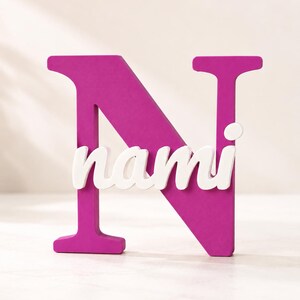 May include: A large, magenta-colored capital letter N made of a solid material. Overlaid across the center of the letter N is the name nami written in a white, lowercase, cursive script font that stands out against the dark pink background.