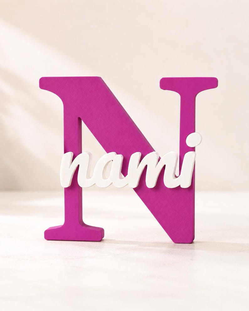 May include: A large, magenta-colored capital letter N made of a solid material. Overlaid across the center of the letter N is the name nami written in a white, lowercase, cursive script font that stands out against the dark pink background.