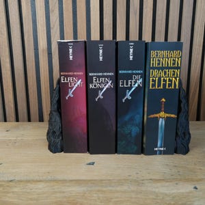 K&ouml;nnte beinhalten: Vier B&uuml;cher mit den Titeln "Elfen Licht", "Elfen K&ouml;nig", "Die Elfen" und "Drachen Elfen" werden gezeigt. Die B&uuml;cher werden von schwarzen Buchst&uuml;tzen gehalten. Die Buchcover sind in verschiedenen Farben gehalten, darunter Rot, Schwarz und Blau.