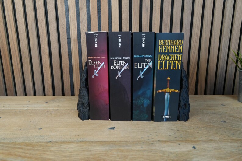 K&ouml;nnte beinhalten: Vier B&uuml;cher mit den Titeln "Elfen Licht", "Elfen K&ouml;nig", "Die Elfen" und "Drachen Elfen" werden gezeigt. Die B&uuml;cher werden von schwarzen Buchst&uuml;tzen gehalten. Die Buchcover sind in verschiedenen Farben gehalten, darunter Rot, Schwarz und Blau.
