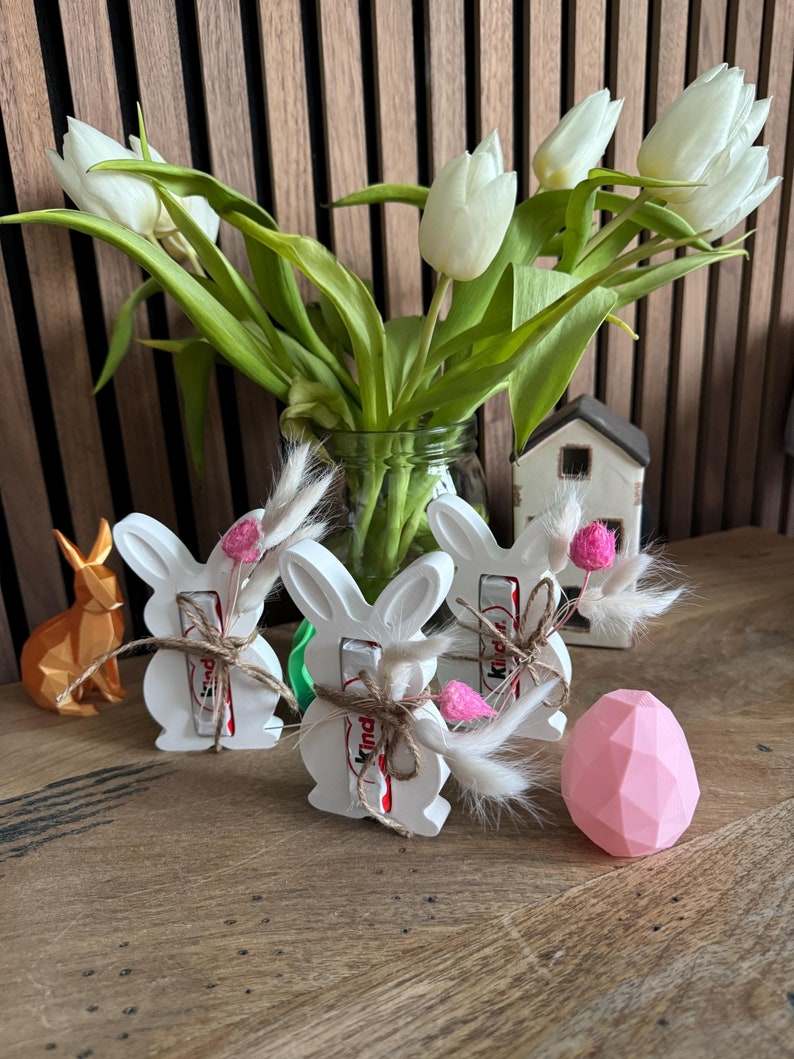 May include: Three white bunny-shaped ceramic holders with a brown and red chocolate bar inside each. The holders are decorated with a brown twine bow and a small bouquet of dried flowers. A pink geometric egg sits on a wooden table in front of the holders.