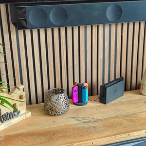 May include: A wooden console table displays a decorative home sign, a metal candle holder, a charging station with colourful game controllers, and decorative ceramic houses. The background features a wooden slatted wall.