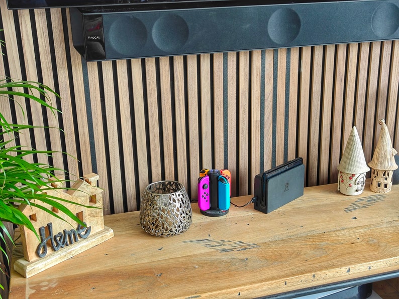 May include: A wooden console table displays a decorative home sign, a metal candle holder, a charging station with colourful game controllers, and decorative ceramic houses. The background features a wooden slatted wall.