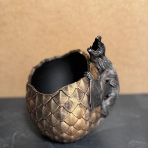 May include: A decorative dragon egg-shaped planter with a detailed scale pattern in gold and bronze tones. A dragon figure is perched on the side with its mouth open. The interior of the planter is black.