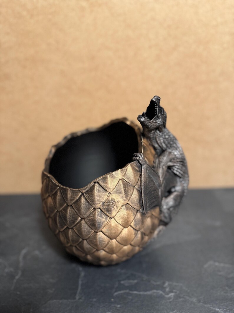May include: A decorative dragon egg-shaped planter with a detailed scale pattern in gold and bronze tones. A dragon figure is perched on the side with its mouth open. The interior of the planter is black.