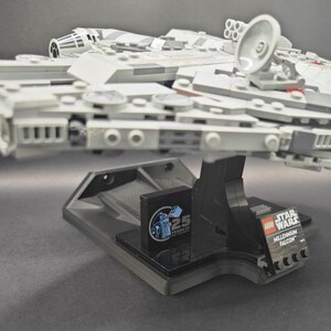 以下が含まれることがあります： 黒いディスプレイスタンドに置かれたグレーのLEGOミレニアム・ファルコン宇宙船。LEGOスターウォーズのロゴと25周年記念のロゴが貼られています。