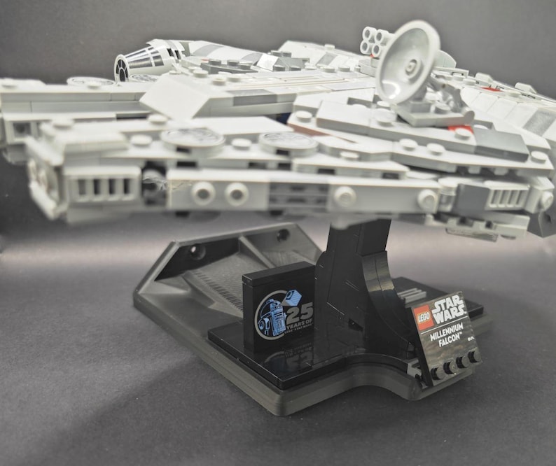 以下が含まれることがあります： 黒いディスプレイスタンドに置かれたグレーのLEGOミレニアム・ファルコン宇宙船。LEGOスターウォーズのロゴと25周年記念のロゴが貼られています。
