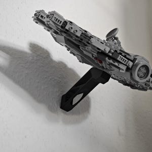 May include: A grey and black model spaceship, mounted on a black stand, appears to be floating against a white wall. The model has intricate details, including a circular design on the front and a radar dish. The shadow of the model is visible on the wall.