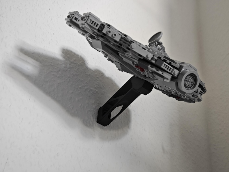 May include: A grey and black model spaceship, mounted on a black stand, appears to be floating against a white wall. The model has intricate details, including a circular design on the front and a radar dish. The shadow of the model is visible on the wall.