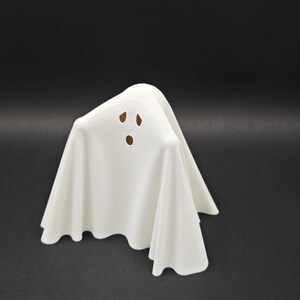 Puede incluir: Una figurita de fantasma blanca con una cara simple, con dos ojos y una boca. El fantasma est&aacute; de pie sobre una superficie negra.