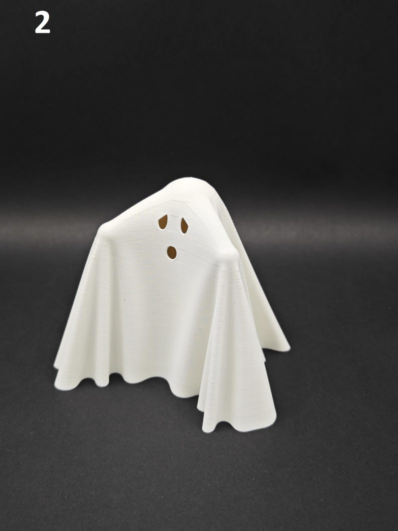 Puede incluir: Una figurita de fantasma blanca con una cara simple, con dos ojos y una boca. El fantasma est&aacute; de pie sobre una superficie negra.