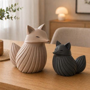 K&ouml;nnte beinhalten: Zwei dekorative Fuchsstatuen. Eine ist hellbeige, die andere schwarz. Beide haben ein stilisiertes Design mit einem strukturierten, gerippten K&ouml;rper und einem spitzen Schwanz. Sie stehen auf einem Holztisch.